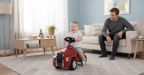 A smiling toddler rides a red toy truck on a living room rug while a man sits on the couch watching him.