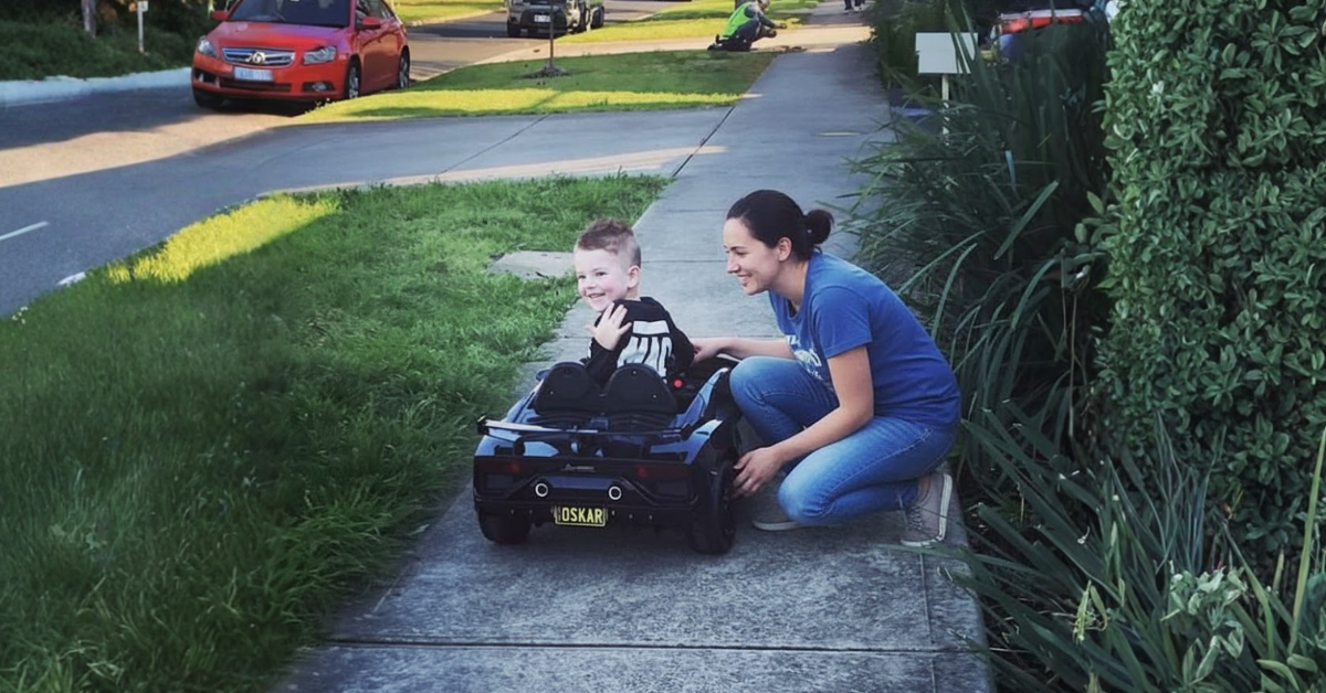 A happy child sits in a black toy car on a sidewalk with a smiling adult crouching beside them, on a sunny day.
