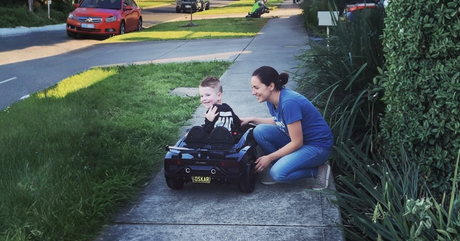 A happy child sits in a black toy car on a sidewalk with a smiling adult crouching beside them, on a sunny day.