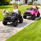 Two children driving ride-on toy off-road vehicles on a concrete path in a backyard, with a small dog running alongside the black vehicle and a pink vehicle behind it. A house and green lawn are visible in the background.