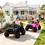 Two children drive toy off-road vehicles on a concrete driveway in front of a white house. The vehicle in the foreground is black and driven by a smiling boy wearing a party hat, while a girl in a pink vehicle drives behind him.