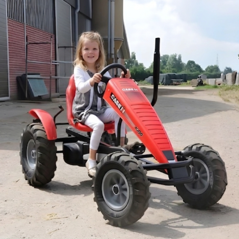 A young girl drives a red Case IH pedal kart on a farm path. It features rugged tractor tires and a vertical exhaust pipe.
