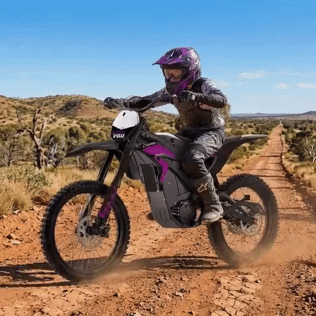A person riding an electric dirt bike on a dusty red dirt road in a dry, scrub-covered landscape under a blue sky. The rider is wearing a purple and black helmet and protective gear, and the bike has black and purple body panels.