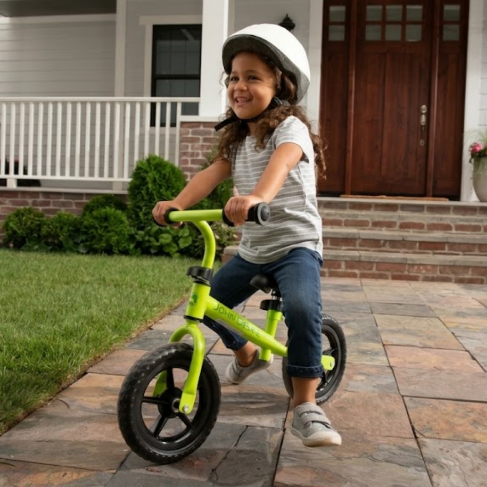 A young girl wearing a white helmet smiles while riding a bright lime green balance bike on a stone patio in front of a house.