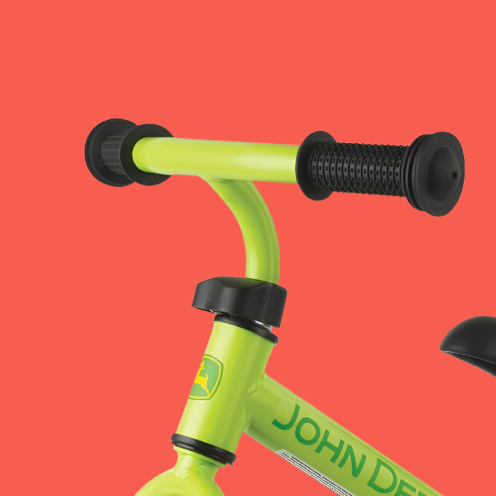 Close-up of a bright lime green balance bike handlebar with black grips and black seat post, featuring the John Deere logo and brand name on the frame, set against a solid coral red background.