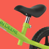 Close-up of a bright lime green balance bike frame with a black saddle and black tire, shown against a solid coral-red background. The brand name 'John Deere' is printed in green on the frame.