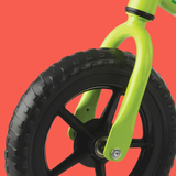 Close-up of a balance bike wheel with a black tire and black spoked wheel, attached to a bright lime green frame against a solid red-orange background.