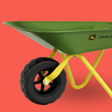 A green wheelbarrow with a yellow frame and black tire, shown against a solid coral-red background. The green metal bed features the John Deere logo.