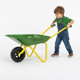 A young boy wearing a green t-shirt and blue denim overalls pushes a green and yellow John Deere wheelbarrow on a clean white background.
