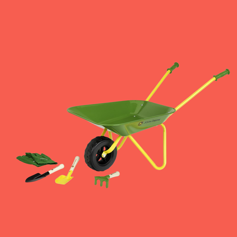 A green and yellow toy wheelbarrow with a black wheel, shown with small gardening tools including a green leaf rake, black-handled trowel, yellow-handled shovel, and green hand fork, on a solid coral-red background.