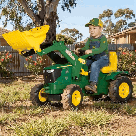 Child playing with a green toy tractor with yellow attachments in a backyard setting.