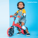 A smiling toddler wears a blue helmet and pads while riding a blue Yvolution balance bike.
