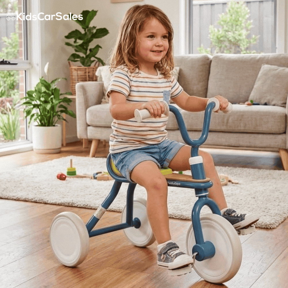 A young child in a striped shirt sits on a blue and tan Yvolution Velo Trike in a living room, looking off-camera and smiling.
