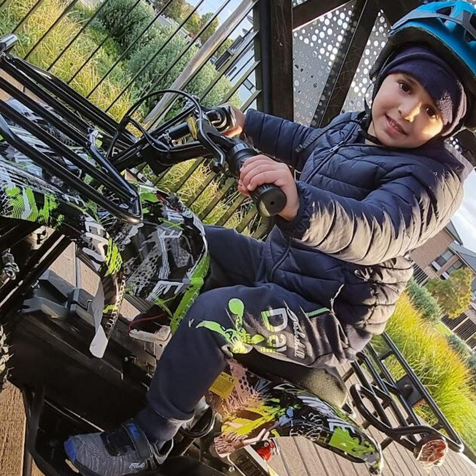 A child wearing a helmet and winter jacket sits on a colourful quad bike, ready to ride under a sheltered area, with a residential neighbourhood and clear sky in the background