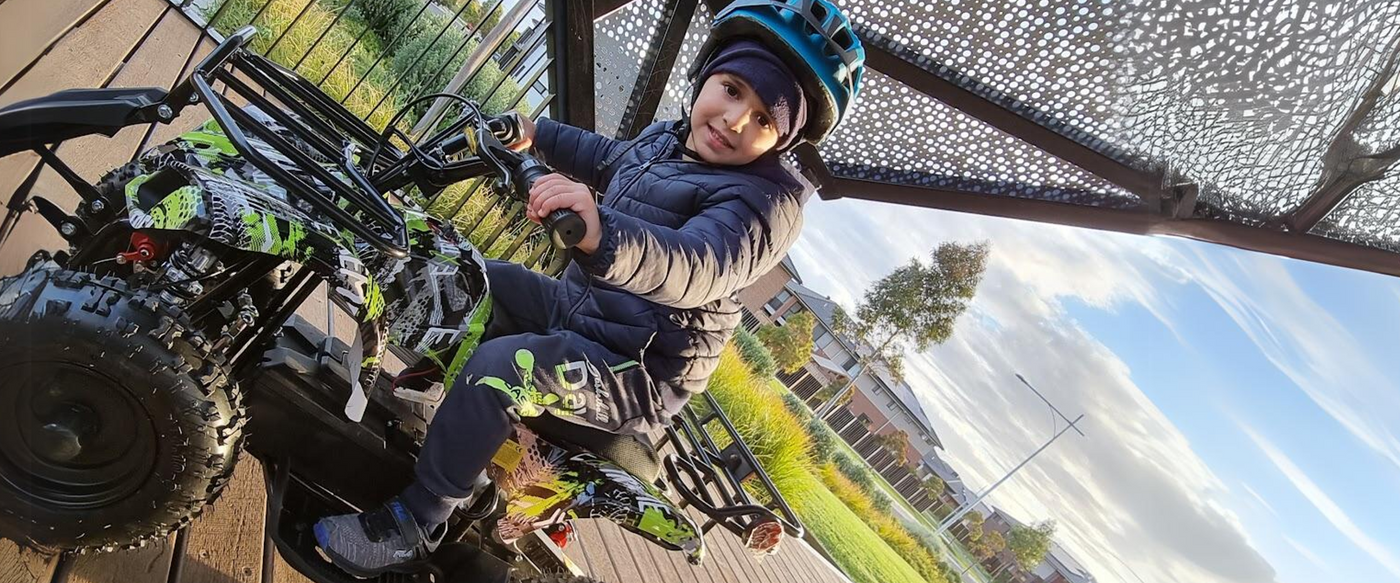 A child wearing a helmet and winter jacket sits on a colourful quad bike, ready to ride under a sheltered area, with a residential neighbourhood and clear sky in the background