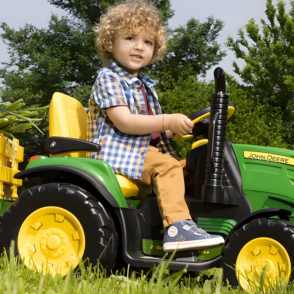 A young child with curly hair riding a green and yellow John Deere toy tractor, pulling a trailer filled with fresh vegetables, in a grassy outdoor setting