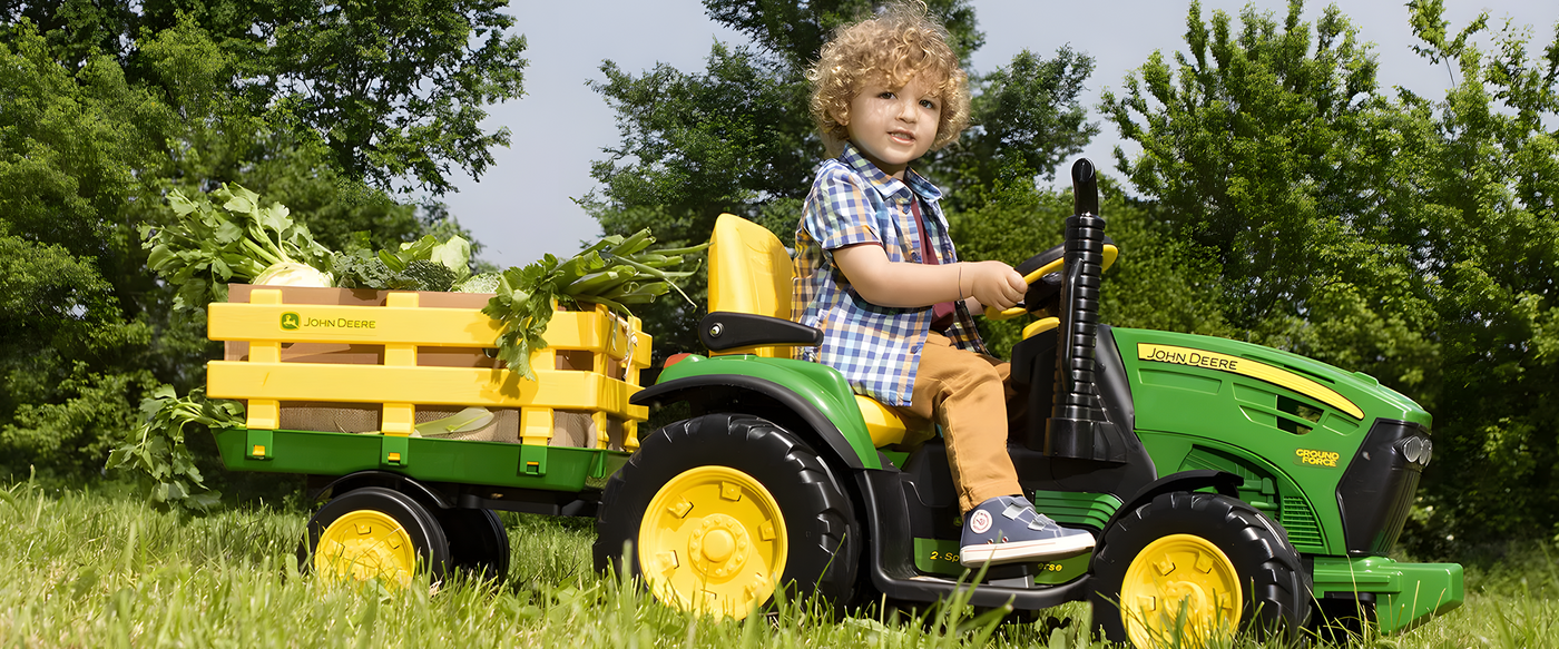 A young child with curly hair riding a green and yellow John Deere toy tractor, pulling a trailer filled with fresh vegetables, in a grassy outdoor setting