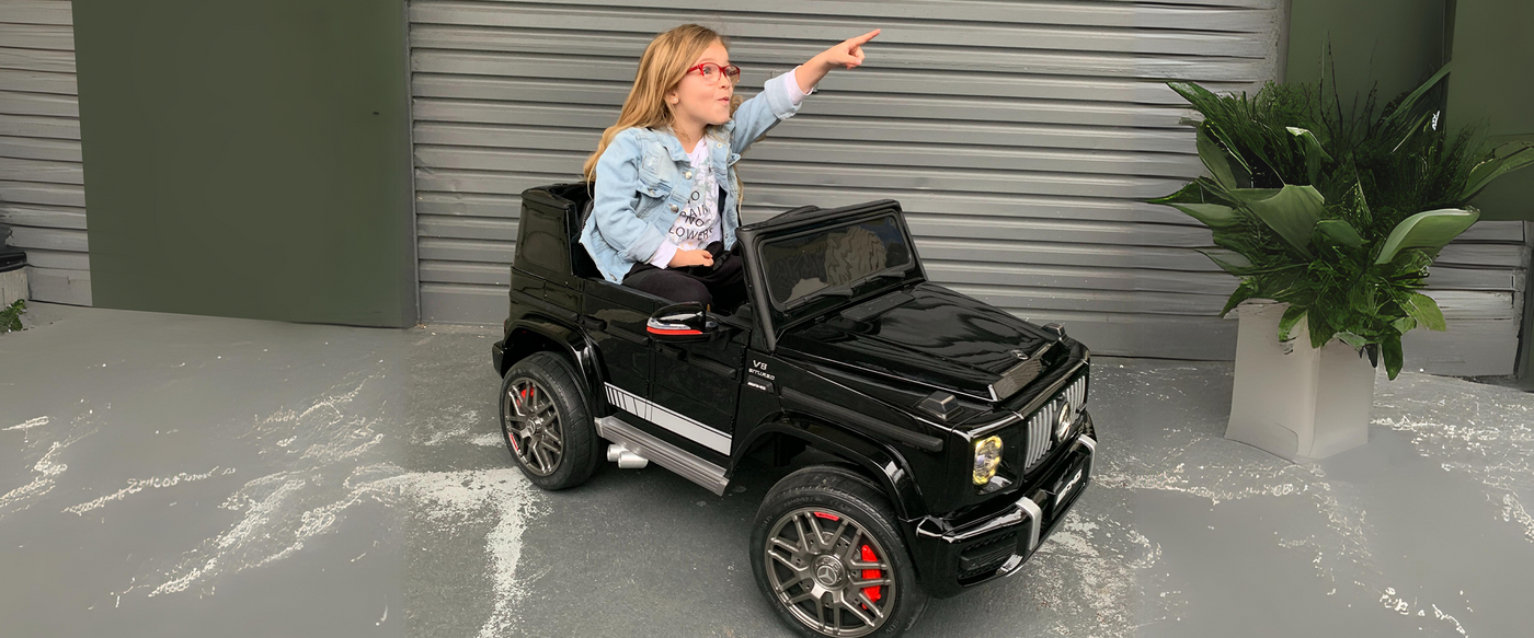 The image shows a young girl wearing a light denim jacket and glasses, joyfully pointing forward while seated in a black ride-on toy car styled after a luxury SUV. The setting is an outdoor space with a modern backdrop, featuring a metal panelled wall and decorative potted plants on either side, creating an urban play environment.