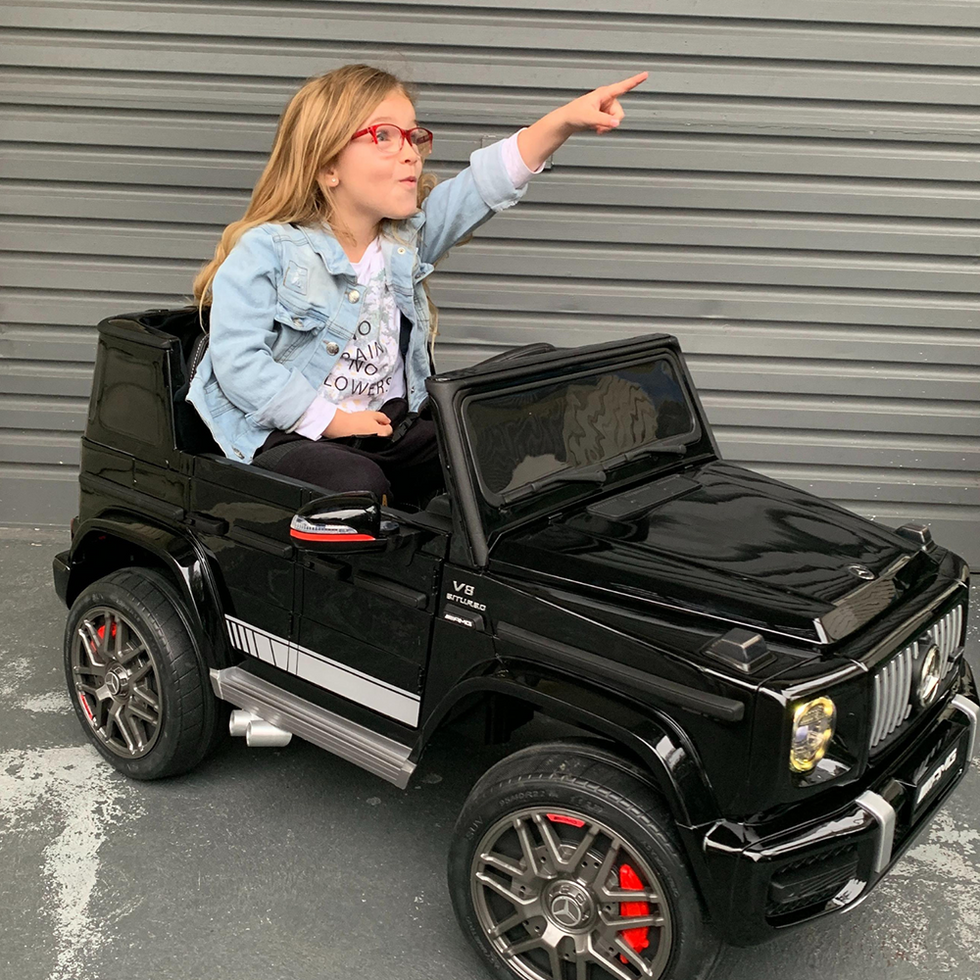 The image shows a young girl wearing a light denim jacket and glasses, joyfully pointing forward while seated in a black ride-on toy car styled after a luxury SUV. The setting is an outdoor space with a modern backdrop, featuring a metal panelled wall and decorative potted plants on either side, creating an urban play environment.