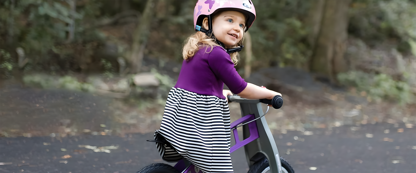 A young girl wearing a pink helmet and a striped dress rides a purple balance bike on a shaded path, with trees in the background