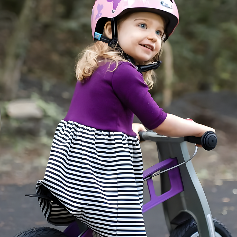 A young girl wearing a pink helmet and a striped dress rides a purple balance bike on a shaded path, with trees in the background