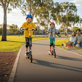 Two kids ride black electric scooters in a park, passing a playground and their waving parents.