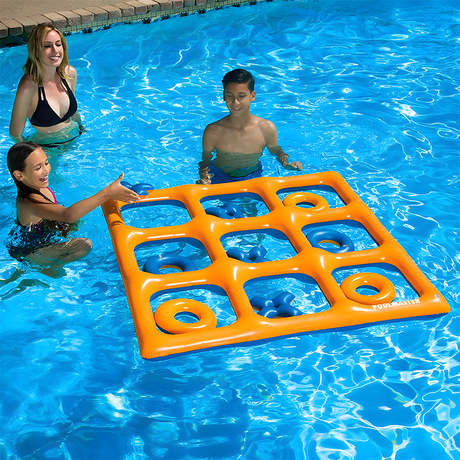 Children playing with an inflatable Tic-Tac-Toe game in a pool. The orange grid floats on the water, with blue 'X' pieces and orange 'O' pieces placed within the grid