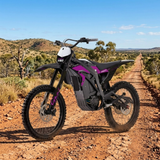 A VEDO electric dirt bike with black and purple body panels is parked on a dirt road in an outdoor desert landscape under a blue sky.