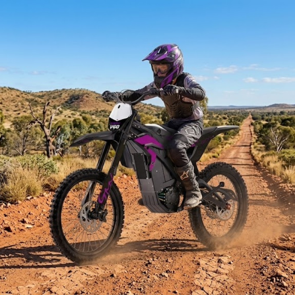 A person riding an electric dirt bike on a dusty red dirt road in a dry, scrub-covered landscape under a blue sky. The rider is wearing a purple and black helmet and protective gear, and the bike has black and purple body panels.