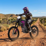 A person riding an electric dirt bike on a dusty red dirt road in a dry, scrub-covered landscape under a blue sky. The rider is wearing a purple and black helmet and protective gear, and the bike has black and purple body panels.