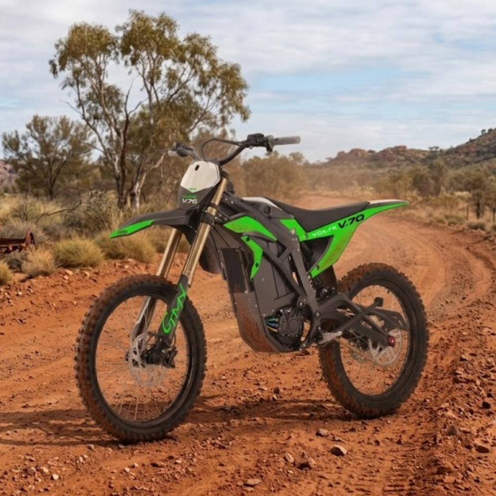 A green and black electric dirt bike with a white front number plate is parked on a reddish-brown dirt road in an outdoor desert landscape with trees and rocky hills under a cloudy sky.