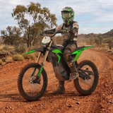 A person in full off-road motocross gear rides a green and black FXR dirt bike on a red dirt trail in a desert landscape.