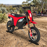 A red and black electric dirt bike with flame graphics and racing decals, parked on a dirt track with trees and spectators in the background.
