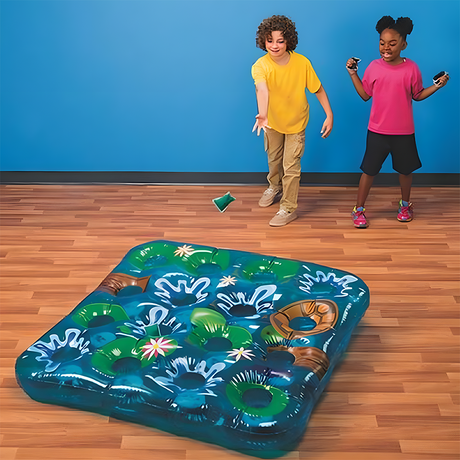 Two children playing an inflatable beanbag toss game with a frog pond theme indoors. One child throws a green beanbag toward the inflatable game board on the wooden floor, which features blue water, frogs, and lily pads