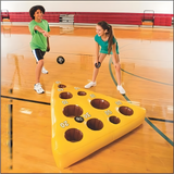 Children playing the Mouse and Cheese Toss game in a gym, trying to throw beanbags into holes on a yellow inflatable cheese mat