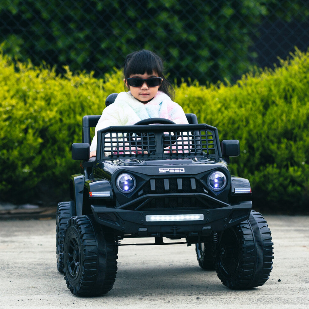 A young girl wearing sunglasses, driving the black Jeep-inspired remote-controlled ride-on electric car, with greenery in the background.