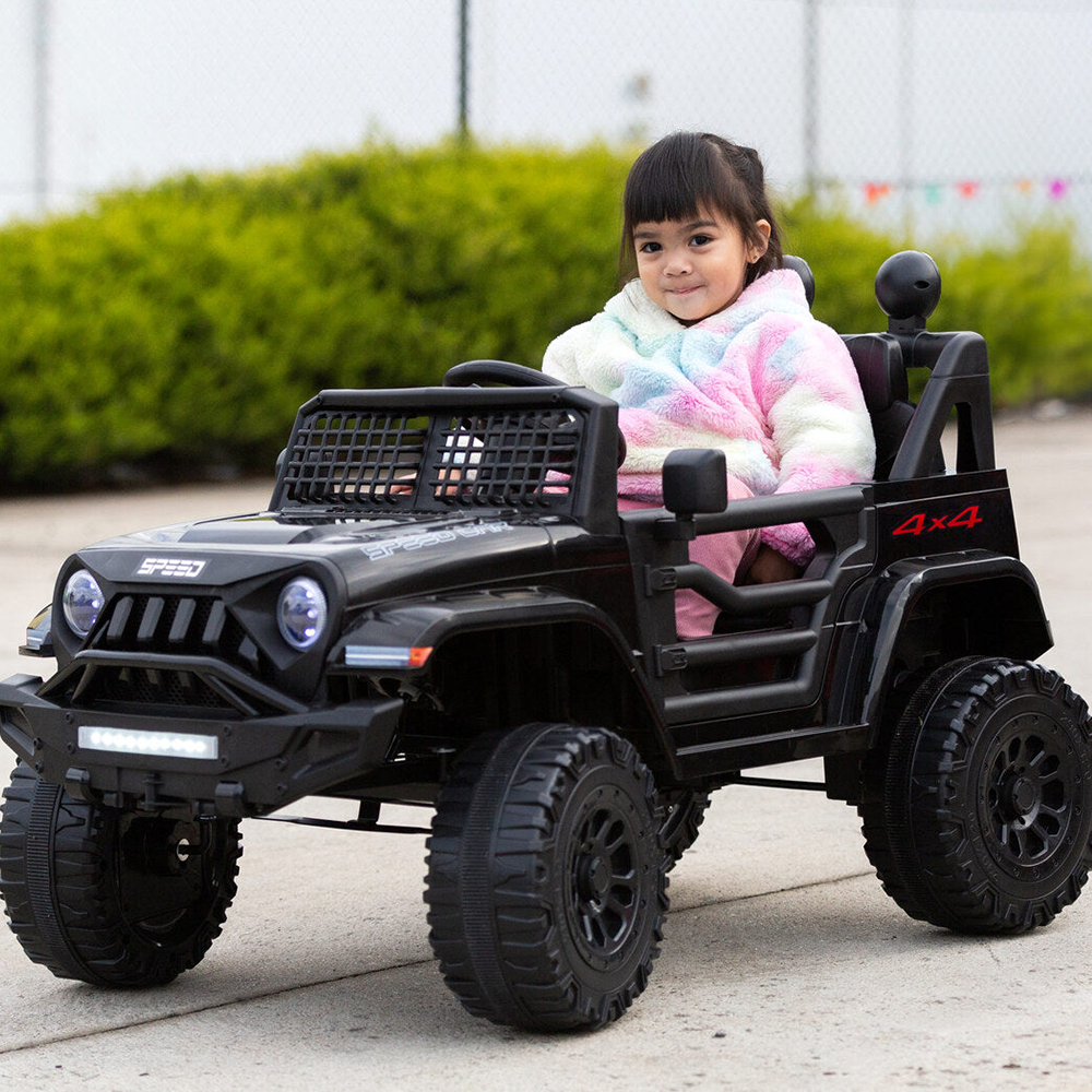 A young girl wearing a colourful jacket, smiling while sitting in a black Jeep-inspired remote-controlled ride-on electric car, outdoors.