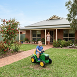 Child on a toy lawn mower in front of a house with a woman standing nearby.