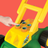 A close-up of a yellow and green toy tractor being pushed by a child's hand, with four colorful buttons on the front panel showing farm-themed icons.