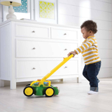 A toddler in a yellow and white striped shirt pushes a green and yellow toy lawnmower in a bright, modern room with a white dresser and framed art.