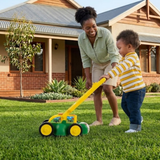 A woman and a young child pushing a green and yellow toy lawn mower on a green grassy lawn in front of a suburban house.