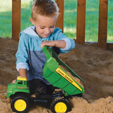A young boy playing in a sandbox with a green John Deere toy dump truck, tipping the bed to unload sand. The boy is wearing a blue and gray hoodie, and the background shows a wooden fence and green grass.