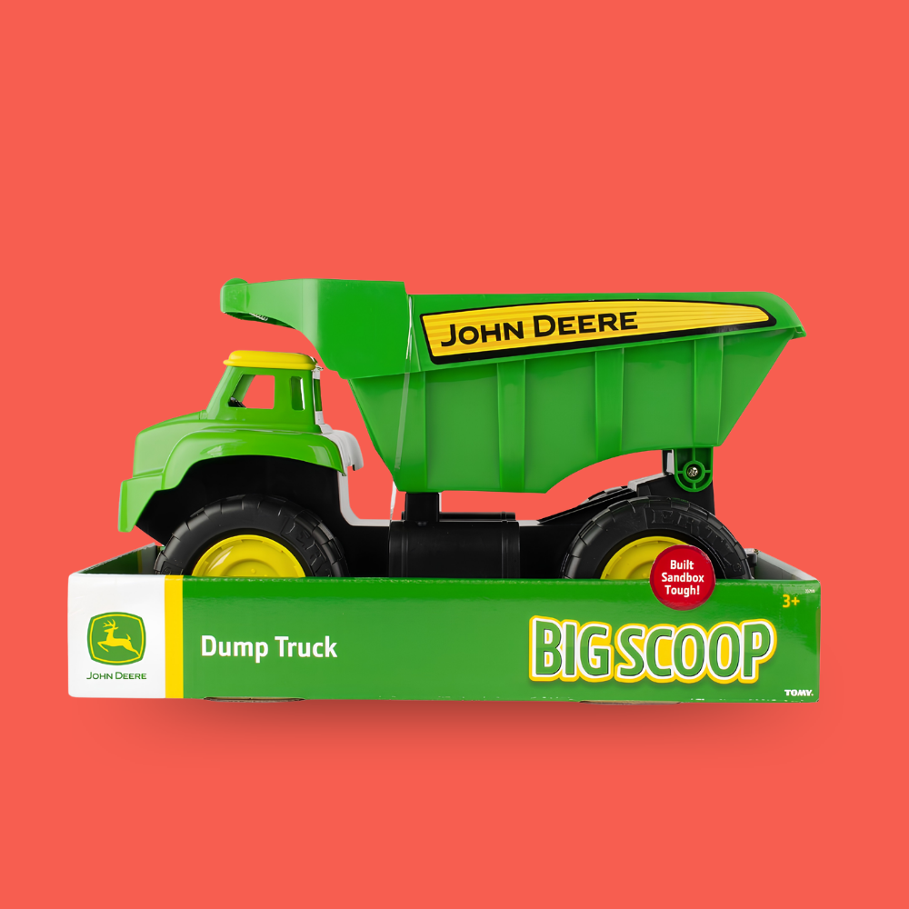 A John Deere dump truck toy in its retail box, set against a solid coral-red background. The green and yellow truck is shown from a three-quarter angle, resting on top of its green and white packaging box which features the text 'John Deere Dump Truck BIG SCOOP'.