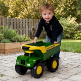 A young child pushing a green and yellow John Deere toy dump truck on a paved backyard path, with a garden, fence, and trees in the background.