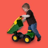 A young child pushing a green and yellow John Deere toy dump truck against a solid coral-red background.