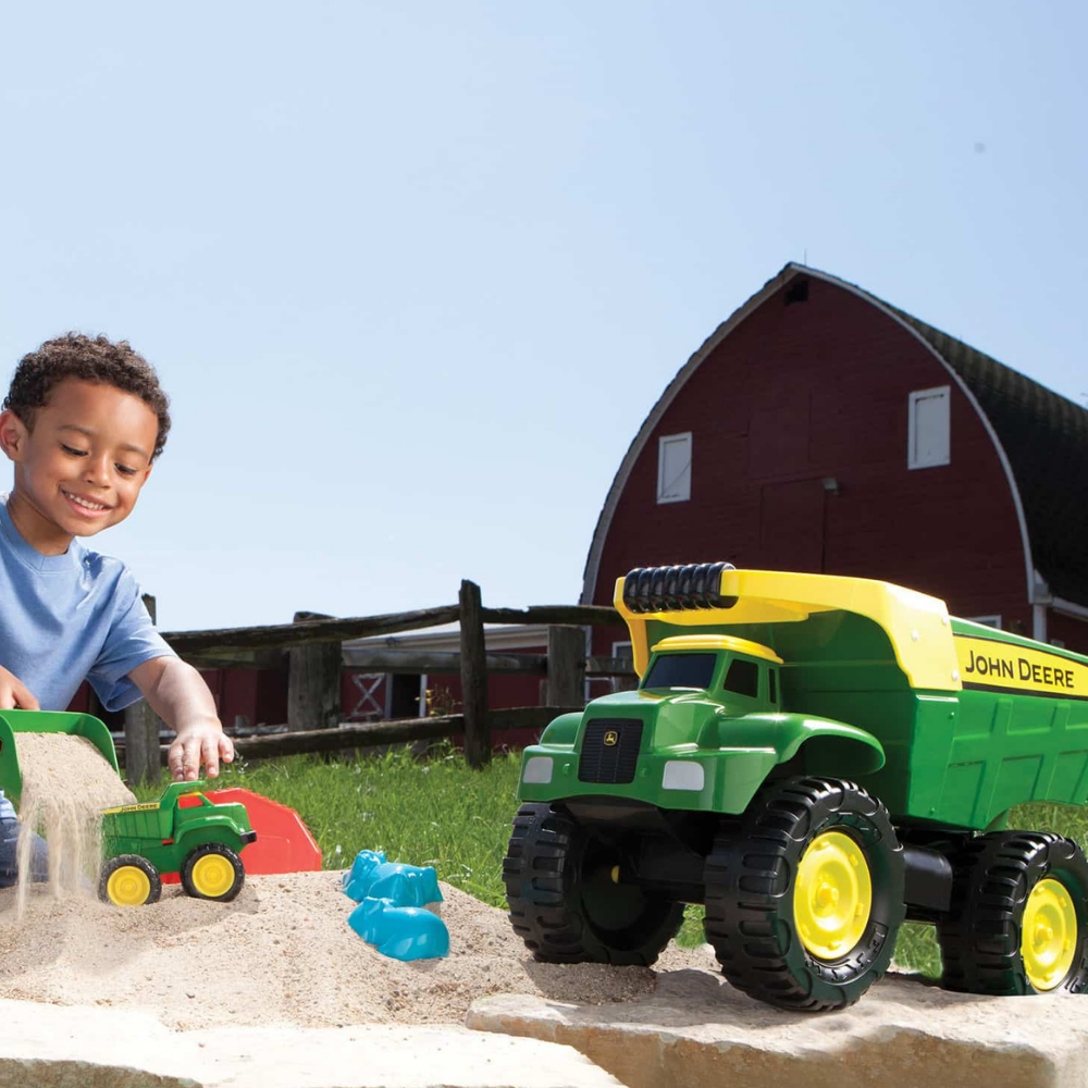 A young boy wearing a blue shirt plays with a large green and yellow John Deere toy dump truck and a smaller toy excavator at a sandbox. In the background, there is a red barn and a blue sky.