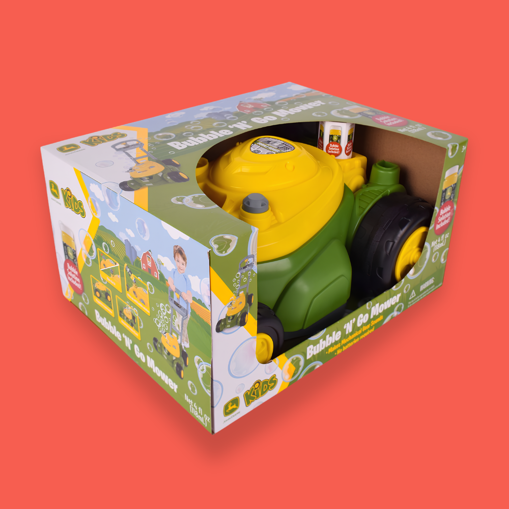 A boxed children's toy lawn mower visible through its retail packaging, photographed on a solid coral-red background. The toy is green with a bright yellow top and black wheels.