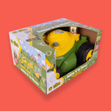 A boxed children's toy lawn mower visible through its retail packaging, photographed on a solid coral-red background. The toy is green with a bright yellow top and black wheels.