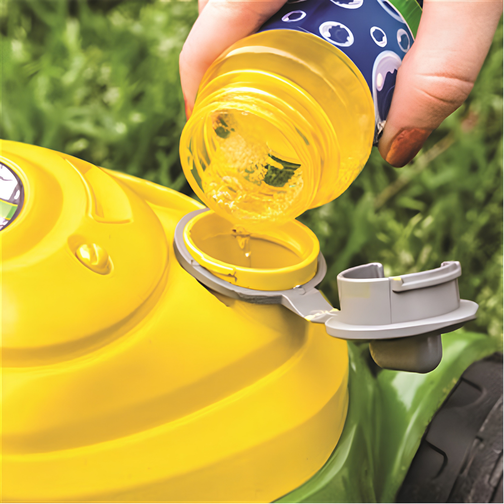 A close-up of a person pouring yellow liquid from a blue bottle into the fuel tank opening of a bright yellow lawn mower outdoors on grass.