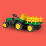 A John Deere toy ride-on tractor with a green and yellow body, black tires, and a matching green and yellow pull-on trailer, shown on a solid coral-red background.